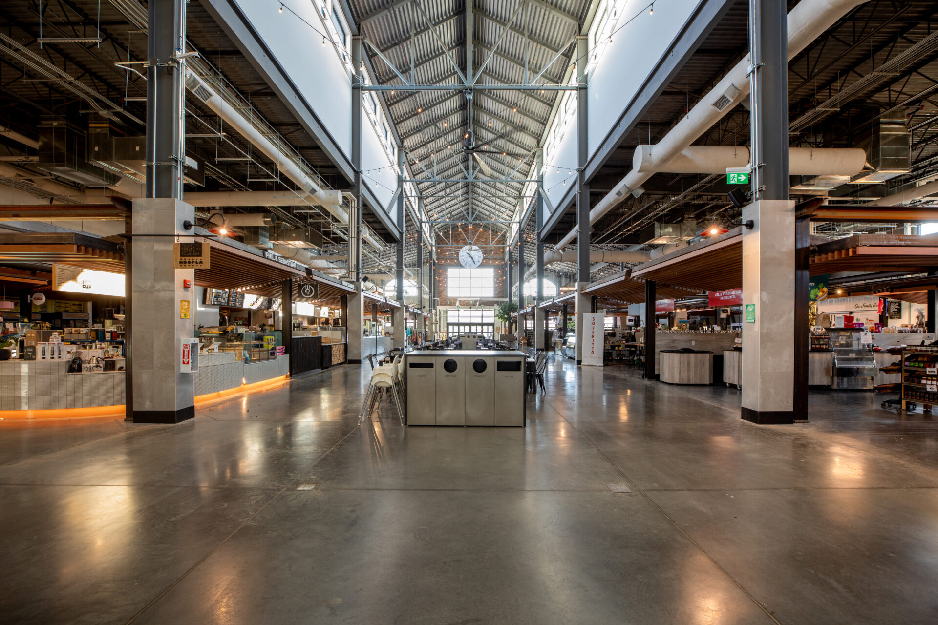 Shot from the centre of the market on the ground level. High glass ceilings surrounded by vendor spaces. Down the centre of the aisle there is seating.